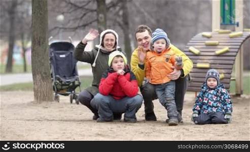 Happy mother, father and their three children waving with the hands. Playground, trees and baby carriage in background.
