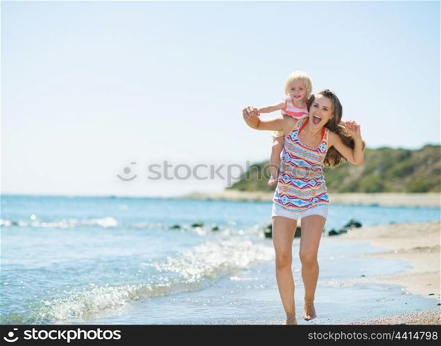 Happy mother holding baby on back and running along sea shore