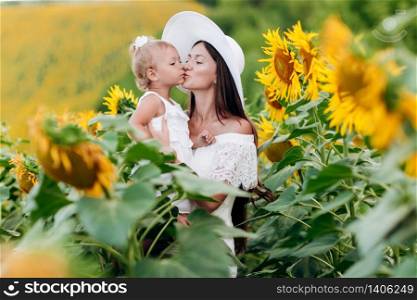 Happy mother in hat with the daughter in the field with sunflowers. mom and baby girl having fun outdoors. family concept. mom kisses her daughter. selective focus.. Happy mother in hat with the daughter in the field with sunflowers. mom and baby girl having fun outdoors. family concept. mom kisses her daughter. selective focus