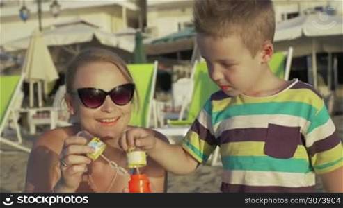 Happy mother in sunglasses and little son blowing bubbles together on the beach. Empty deck-chairs and hotels in background. Having fun with mom