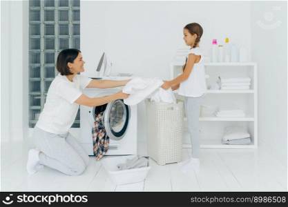 Happy mother loads clothes in washing machine, little girl helps, gives white linen from basket, do housework together, dressed in casual clothing. Daughter offers help to mum, clean clothes
