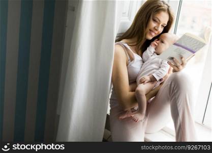 Happy mother reading book  with newborn baby girl by the window