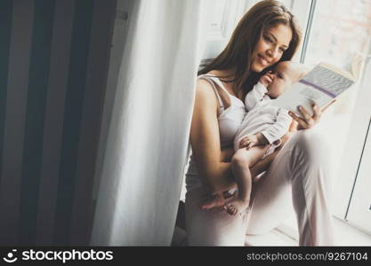 Happy mother reading book with newborn baby girl by the window