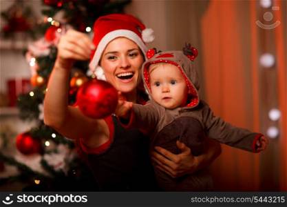 Happy mother showing Christmas ball to baby near Christmas tree&#xA;