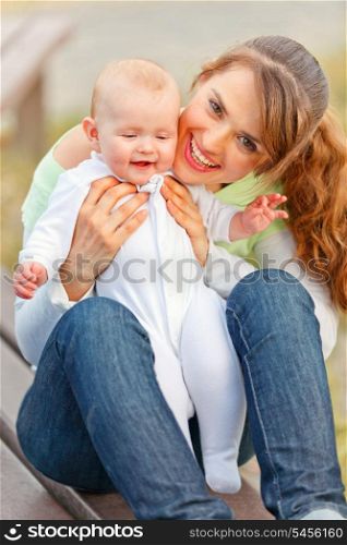 Happy mother sitting on bench in park and holding smiling adorable baby girl in hands&#xA;
