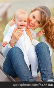 Happy mother sitting on bench in park and holding smiling adorable baby girl in hands&#xA;