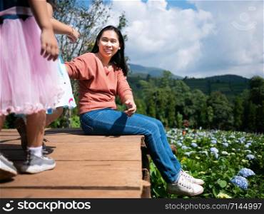 Happy mother sitting with her daughter on wooden ground over trees with mountains background under blue sky in summer day.