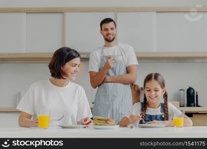 Happy mother talks to daughter while have breakfast. Father stands behind, prepared delicious dish for family. Friendly family members meet at kitchen during weekend, enjoy nice conversation