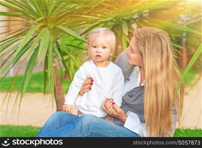 Happy mother with baby girl sitting on green field in tropical park in bright sunny day, enjoying motherhood, love and happiness concept