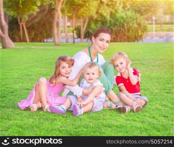 Happy mother with cute little babies sitting on fresh green grass field, having fun outdoors, enjoying parenthood, happiness and love concept