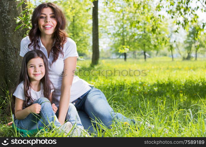 Happy mother with her daughter sitting on grass under the tree in park. Mother with daughter in park