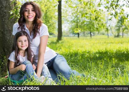 Happy mother with her daughter sitting on grass under the tree in park. Mother with daughter in park