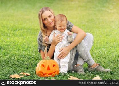 Happy mother with little baby outdoors, sitting on green grass field with orange carved pumpkin, preparation to Halloween holiday