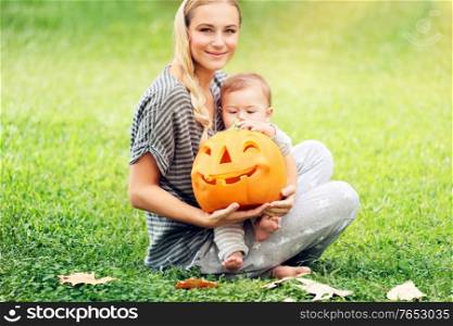 Happy mother with little baby outdoors, sitting on the grass field with orange carved pumpkin toy, preparation to Halloween holiday