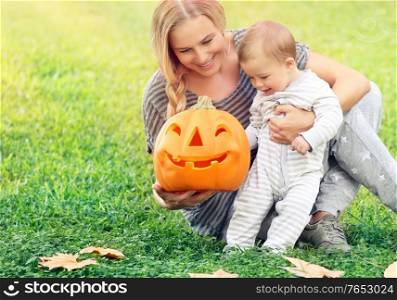 Happy mother with little baby outdoors, sitting on the green grass field and playing with orange carved pumpkin with smiling face,enjoying Halloween holiday