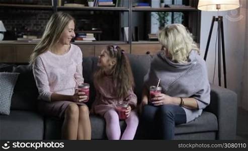Happy multi generation family with mason jars of fresh berry smoothies in hands communicating while sitting on cozy sofa in living room. Cheerful grandmother, mother and sweet daughter spending great time together at home on the couch. Slo mo.
