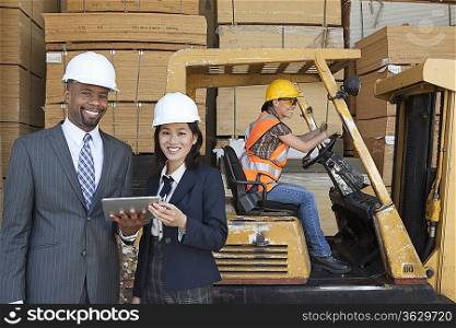 Happy multiethnic engineers holding tablet PC with female worker driving forklift truck in background