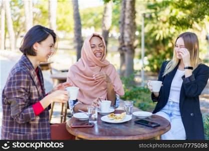 Happy multiethnic female friends laughing while sitting at table with snack and coffee on terrace of cafeteria on summer day. Cheerful multiracial women sitting in outdoor cafe
