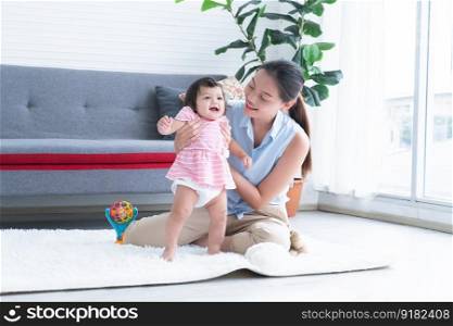 Happy multiracial family, young Asian mother holding and supporting Caucasian newborn 7 months old to stand and walk first step on floor at home with toy. Education and development children concept