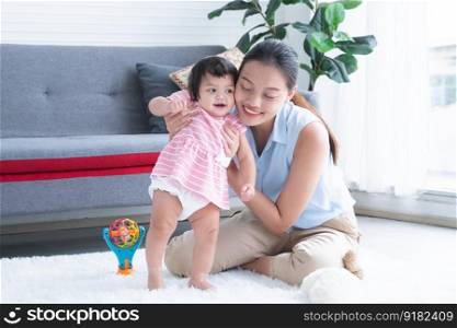 Happy multiracial family, young Asian mother holding and supporting Caucasian newborn 7 months old to stand and walk first step on floor at home with toy. Education and development children concept