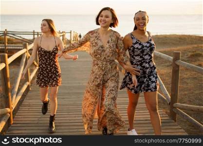 Happy multiracial women holding hands and reaching hand to girlfriend while strolling on wooden walkway on seashore at sunset. Smiling multiethnic girlfriends walking on wooden boardwalk on sea shore