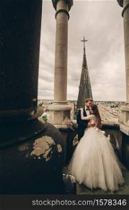 Happy newlywed. beautiful bride and stylish groom are hugging on the balcony of old gothic cathedral with panoramic city views.. Happy newlywed. beautiful bride and stylish groom are hugging on the balcony of old gothic cathedral with panoramic city views