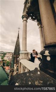Happy newlywed. beautiful bride and stylish groom are kissing on the balcony of old gothic cathedral with panoramic city views.. Happy newlywed. beautiful bride and stylish groom are kissing on the balcony of old gothic cathedral with panoramic city views