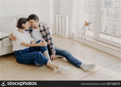 Happy newlywed couple move in new apartment, rejoice having spare time together, embrace and touch noses, going to have sweet kiss, pose on floor in empty room near sofa, their dog in background