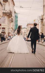 Happy newlyweds couple on a walk in old European town street, gorgeous bride in white wedding dress together with handsome groom. rear view.. Happy newlyweds couple on a walk in old European town street, gorgeous bride in white wedding dress together with handsome groom. rear view