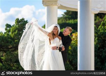 Happy newlyweds hugging on a walk, the girl raised her veil with her hand and smiles