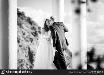 Happy newlyweds kiss in a beautiful gazebo with columns against the background of foliage and sky, black and white version
