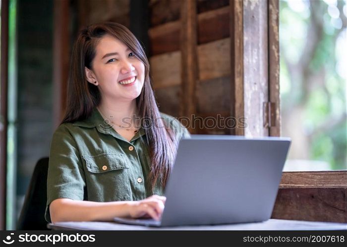 Happy of asian freelance people business female wearing brownish green dress stylish hipster casual working with laptop computer with coffee cup beside the window of a wooden house background