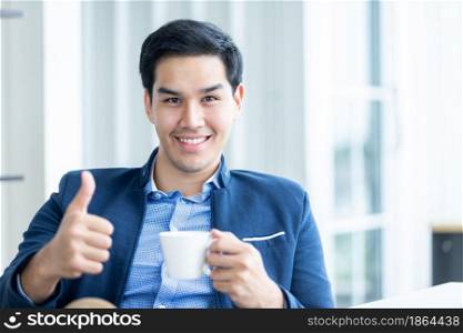 Happy of asian young businessman see a successful business plan on the laptop computer and holding a coffee cup, pen on wooden table background in office,business expressed confidence embolden