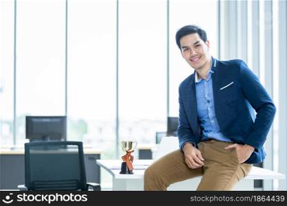 Happy of asian young businessman see a successful of wrist in a business suit of man and wearing blue jacket a champion cup on table at In the office room.