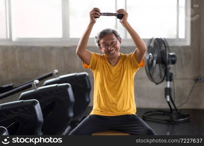 Happy old man exercise dumbbell in modern fitness gym, using sportive equipment. Fit senior guy with gray hair biceps workout. Bodybuilding and elderly Healthy lifestyle.