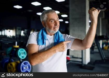 Happy old man shows his muscles, gym interior on background. Sportive grandpa on fitness training in sport center. Healty lifestyle, health care, elderly sportsman. Happy old man shows his muscles in gym