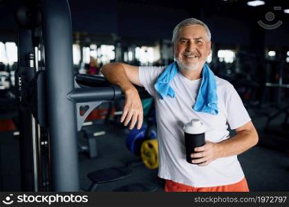 Happy old man with bottle of water and towel, gym interior on background. Sportive grandpa on fitness training in sport center. Healty lifestyle, health care, elderly sportsman. Old man with bottle of water and towel in gym