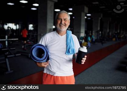 Happy old man with bottle of water, towel and mat, gym interior on background. Sportive grandpa on fitness training in sport center. Healty lifestyle, health care, elderly sportsman. Old man with bottle of water, towel and mat in gym