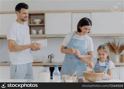 Happy parents cook together with daughter, pose at modern home kitchen give eggs to add to dough, small kid whisks ingredients in bowl have glad expressions. Food and family concept. Cooking breakfast