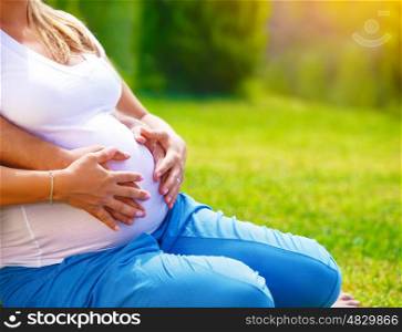Happy parents expecting baby, pregnant woman with husband touching belly, sitting on green grass, body part, young family and new life concept