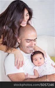 Happy parents with adorable little baby girl sitting on the couch at home, enjoying parenthood, young happy family, new life concept