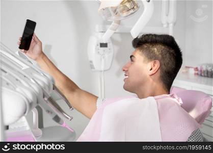 Happy patient taking selfie at dental clinic. Patient is holding smartphone and showing his healthy smile. High quality photo.. Happy patient taking selfie at dental clinic. Patient is holding smartphone and showing his healthy smile.