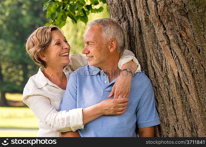 Happy pensioner couple cuddling outdoors leaning against tree