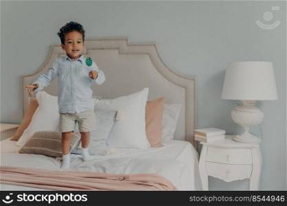 Happy playful afro american child boy with lollipop having lots of fun in bedroom, jumping on bed mattress with variety of pillows and eating candy while mom is not watching. Childhood concept. Carefree cute afro american child boy with lollipop jumping ob bed at home