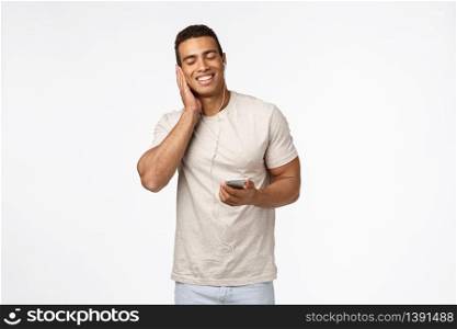 Happy, pleased handsome man enjoy listening favorite music, holding smartphone, press earphone to ear and close eyes, smiling delighted, satisfied with fantastic headphones quality, white background.. Happy, pleased handsome man enjoy listening favorite music, holding smartphone, press earphone to ear and close eyes, smiling delighted, satisfied with fantastic headphones quality, white background