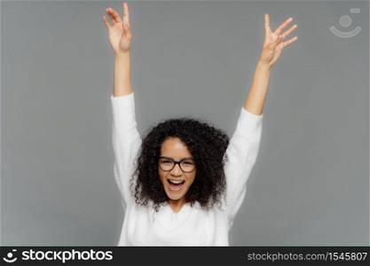 Happy pleased young woman raises hands up, being in high spirit, dances over grey background, celebrates someting, has Afro haircut, wears white jumper, transparent glasses for vision correction