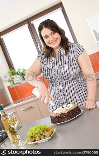 Happy plus size woman with white wine, chocolate cake and cream in modern kitchen