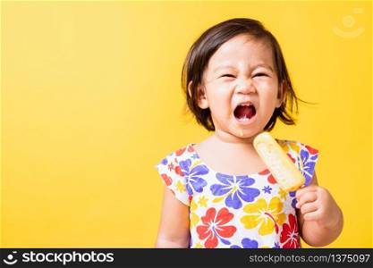 Happy portrait Asian baby or kid cute little girl attractive laugh smile wearing dick pattern shirt holds and eating sweet wooden ice cream, studio shot isolated on yellow background, summer concept
