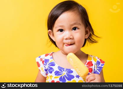 Happy portrait Asian baby or kid cute little girl attractive laugh smile wearing dick pattern shirt holds and eating sweet wooden ice cream, studio shot isolated on yellow background, summer concept