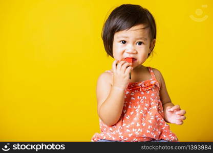 Happy portrait Asian baby or kid cute little girl attractive laugh smile wearing t-shirt playing holds cut watermelon fresh for eating, studio shot isolated on yellow background, healthy food and summer concept